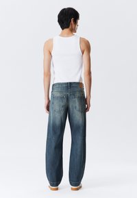 Mies, joka on pukeutunut valkoiseen hihattomaan paitaan ja löysiin sinisiin farkkuihin, seisoo selin yksinkertaista valkoista taustaa vasten.