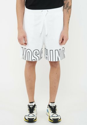 Man draagt witte short met trekkoord en grote "MOSCHINO" tekst aan de voorkant, en witte EA7 sneakers met zwarte veters.