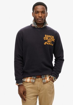 Navy sweatshirt met oranje grafische tekst. Gelaagd geruit overhemd in blauw, geel en rood. Beige broek. Zilveren horloge zichtbaar om de pols.