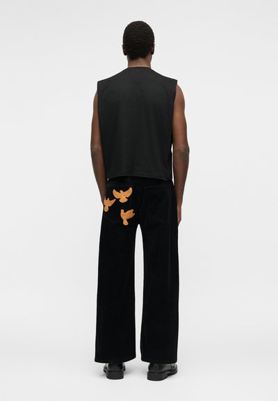 Άνδρας που φοράει μαύρο αμάνικο τοπ και μαύρο παντελόνι με φαρδιά παντελόνια και τρεις πορτοκαλί διακοσμήσεις πουλιών στην πίσω τσέπη, στέκεται μπροστά σε μονόχρωμο φόντο.