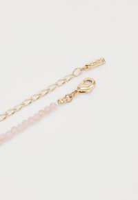 Gullfarget kjede med enkel lås, med rosa fasetterte perler og et merket tag. Glatt metall og polerte steiner.