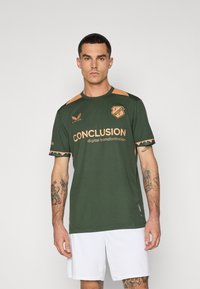 Zaļš sporta krekls ar oranžiem akcentiem. Priekšpusē ir uzraksts "SECINĀJUMS" un logo. Īsas piedurknes, apaļa apkakle un sintētiska auduma.