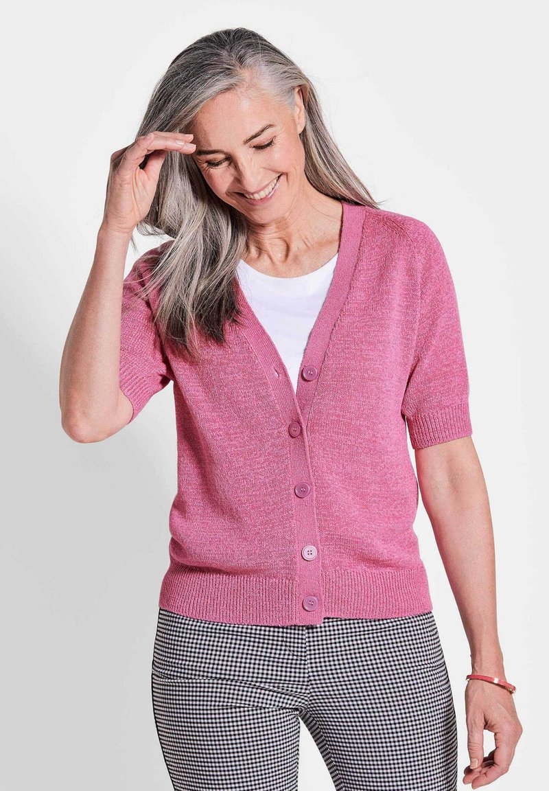 Rosa kurzärmelige Strickjacke mit V-Ausschnitt, Vorderknöpfen, geripptem Saum, getragen über einem weißen Hemd und kombiniert mit schwarz-weißen karierten Hosen.