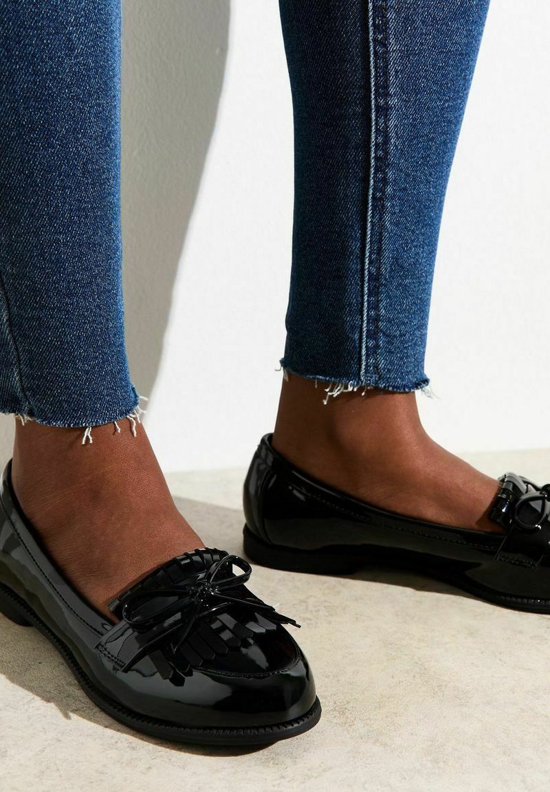 Mocassins en cuir verni noir avec des franges et un détail de nœud. Profil bas et bout arrondi. Associés à un denim usé de longueur cheville.