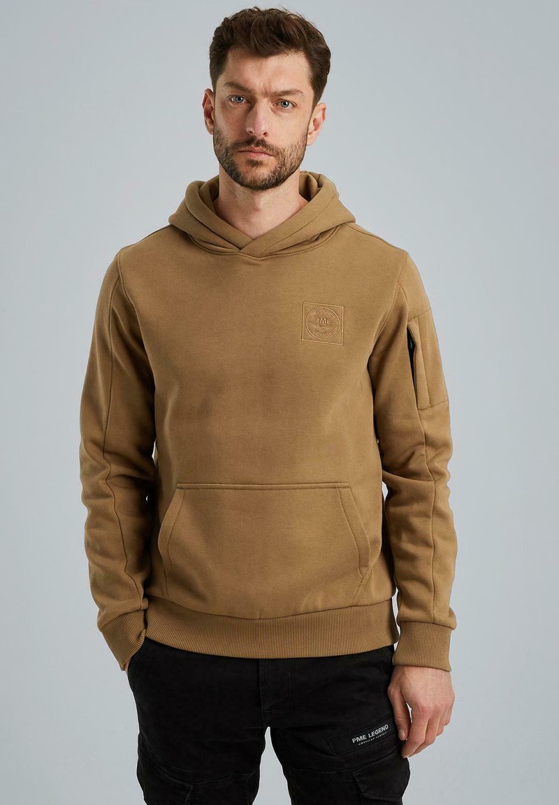 Hooded sweatshirt in light brown cotton blend, with ribbed cuffs, front pocket, and a logo patch on the left chest. Smooth texture.