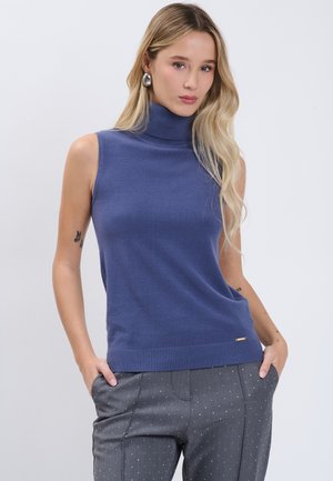 Frau mit langen blonden Haaren trägt einen ärmellosen blauen Rollkragenpullover und graue gepunktete Hose, steht mit den Händen in den Taschen.