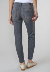 Osoba v upevněných šedých džínách a krémových mokasínách, stojící s jednou rukou u zadní kapsy na jednobarevném pozadí.