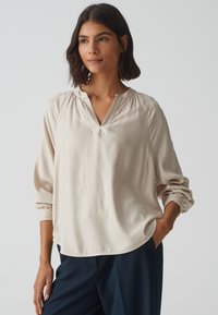 Lichtbeige blouse met lange mouwen en een v-hals, met geplooide details op de schouders en een relaxte pasvorm, gemaakt van een soepele, zachte stof.
