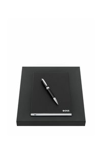 Carnet à couverture rigide noire avec une étiquette "BOSS" et un stylo noir à accents argentés posé dessus, le tout placé sur une boîte rectangulaire noire.