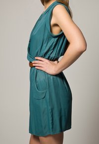 Teal sleeveless dress with a draped design, belted waist, and side pockets. Made of smooth fabric with a lightweight texture.