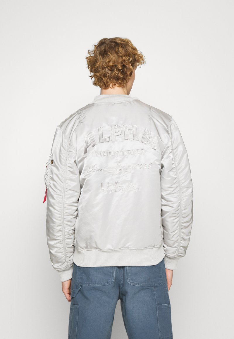 Veste bomber argentée avec un col et des poignets côtelés, comportant un texte brodé au dos. Comprend des poches zippées.