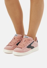 Roze suede sneakers met zwarte accenten, vetersluiting en een witte rubberen zool. Bevat branding op de tong en zijkant.
