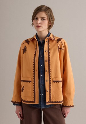Donna che indossa una giacca giallo senape con bordatura floreale e a zigzag marrone scuro sopra una camicia di denim scuro abbottonata e pantaloni marroni.