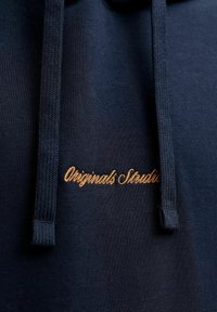 Tume meriinovärvi kapuutsiga pusa, millel on punutud nöörid ja kuldne tikand "Originals Studio" rinnal, kirjutatud kursiivis.