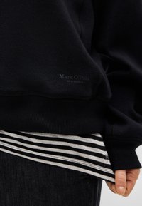 Melns džemperis ar apaļu kakla izgriezumu, tai ir teksturēts audums un smalks logotips. Zem tā redzama baltā un melnā svītraina garām piedurknēm krekls.