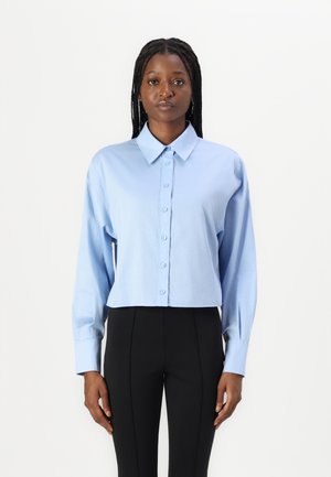 Lys blå cropped skjorte med klassisk krage, knappelukking og lange ermer; stoffet ser glatt ut med en lett glans. Kombinert med svarte bukser.
