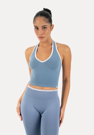 Femme portant un crop top bleu halter assorti et des leggings taille haute avec une bordure blanche, debout devant un fond clair uni.