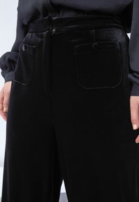 Crne baršunaste hlače imaju visoki struk, dva prednja džepa s kopčanjem, te detalj remena, što stvara glatku, teksturiranu završnicu.