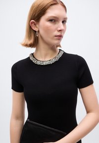 Μαύρο ριμπ φόρεμα με μεταλλικό γιακά, κοντά μανίκια και εφαρμοστή γραμμή. Το μοντέλο κρατάει μαύρο φάκελο, παρουσιάζεται πάνω σε φωτεινό φόντο.