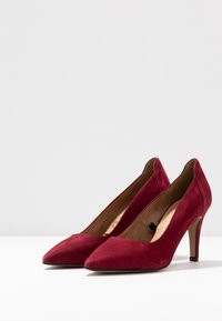 Červené sametové lodičky na vysokém podpatku se špičatou špičkou a 7,5 cm stiletto podpatkem. Hladká textura a minimalistický design s zužující se siluetou.