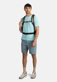 Svetle modré sportovní tričko, šedé šortky a batoh s nastavitelnými popruhy. Tenisky s béžovým a oranžovým designem. Kšiltovka na hlavě.