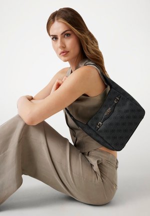 Sac à épaule noir à motif avec des accents métalliques, présentant une texture lisse et une forme structurée, porté avec une tenue de teinte neutre.