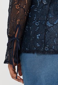 Top in pizzo blu navy con maniche lunghe, caratterizzato da un motivo floreale e nastri legati ai polsini. Il pizzo strutturato Contrasta con il denim liscio.
