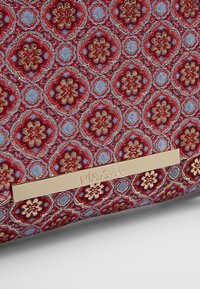 Tissu à motif floral rouge et or avec des accents bleus et une étiquette en métal doré gravée de "MAX&Co." sur une surface texturée.