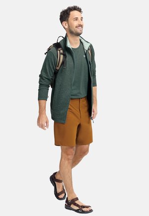 Junger Mann trägt grüne Jacke, grünes T-Shirt, braune Shorts, Sandalen und einen Rucksack, geht lächelnd.