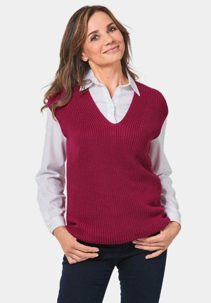 Gilet en tricot bordeaux avec un col en V profond, en tissu texturé, porté sur une chemise blanche à col, associé à un jean sombre, les mains sur les hanches.