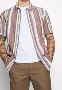 Chemise à manches courtes à boutons, rayée rouge, bleue et beige, réalisée en tissu léger, avec une poche poitrine et une coupe décontractée.