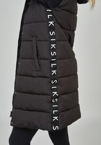 Black puffer vest with quilted texture, featuring white logo tape on the side, and side pockets for convenience. Zipper closure.