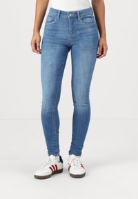 Tynne blå denimjeans med høy midje, fem lommer og subtile sømdetaljer. Brukt med hvite sneakers med røde detaljer.