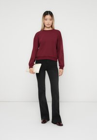 Kvinna som står mot vit bakgrund, iklädd en vinröd sweatshirt, svarta utsvängda byxor, vinröda skor och håller en vit clutchväska i vänster hand.