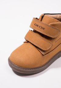 Braune Wildleder-Ankle-Boots mit zwei verstellbaren Klettverschlüssen, wasserdichten Eigenschaften und Gummisohle, die Nähte und Logo zeigt.