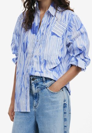 Žena v volné modro-bíle pruhované košili na knoflíky s vyhrnutými rukávy a světle modrých džínech s vysokým pasem, ruka v kapse.