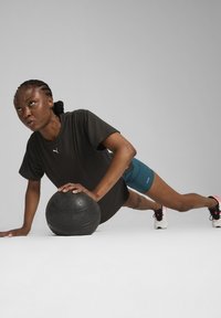 Zwarte kortemouwenshirt, teal korte broek en witte sportschoenen met roze accenten. Gebruiker in plankpositie met een gestructureerde zwarte medicijnbal.
