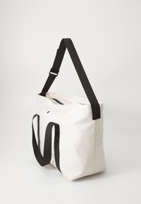 Bolso tote blanco hecho de tela duradera, con asas negras que llevan el logo, gran compartimento principal, cierre superior con cremallera y bolsillo lateral.