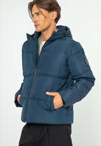 Marineblauwe pufferjack met capuchon, ritssluiting aan de voorkant, zijzakken en stiksels. Glad textuur; logo patch op de mouw.