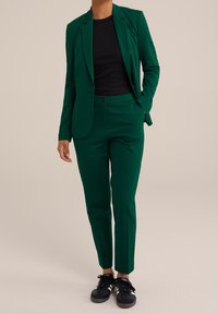 Person klädd i en mörkgrön kostym med svart skjorta och svarta sneakers med vita ränder, stående mot en enkel bakgrund.