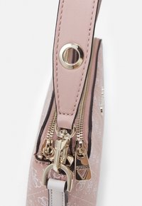 Nahaufnahme eines rosa gemusterten Handtaschenriemens mit goldenen Reißverschluss-Ziehlingen, von denen einer mit dem "GUESS"-Logo graviert ist.