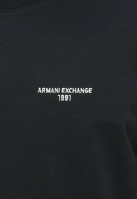 Melns kokvilnas T-krekls ar apaļu kakla izgriezumu, kam vidū krūtīs ir balts izšūts teksta "ARMANI EXCHANGE 1991".