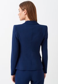 Blazer sartorial blu navy con silhouette aderente, revers a scacco, maniche lunghe e una texture del tessuto liscia.