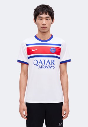 Jeune homme portant un maillot blanc du Paris Saint-Germain avec des bandes rouges et bleues et le logo Qatar Airways, debout devant un fond uni.