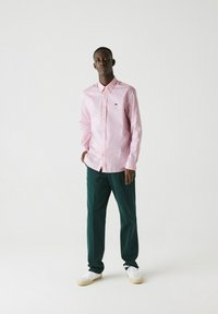 Camicia a maniche lunghe rosa chiaro con colletto, abbinata a pantaloni verde scuro. Le sneakers bianche completano l'outfit.