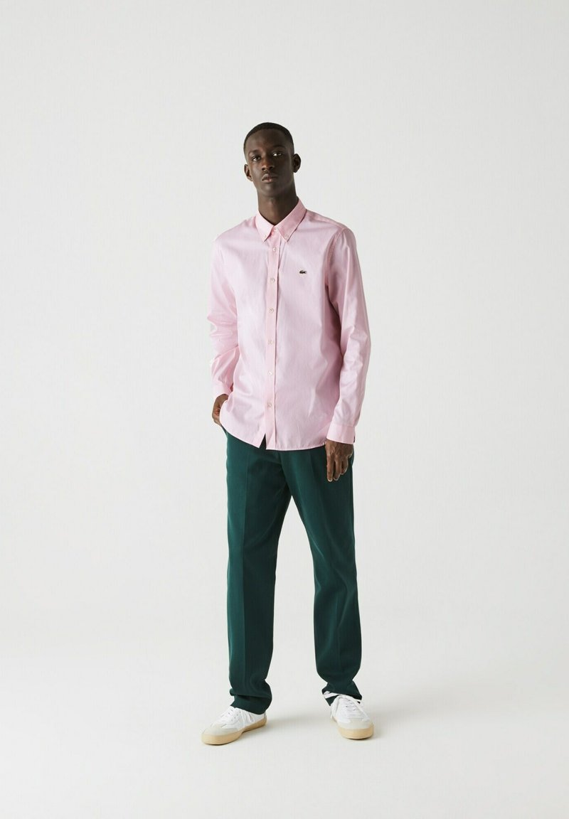 Camicia a maniche lunghe rosa chiaro con colletto, abbinata a pantaloni verde scuro. Le sneakers bianche completano l'outfit.
