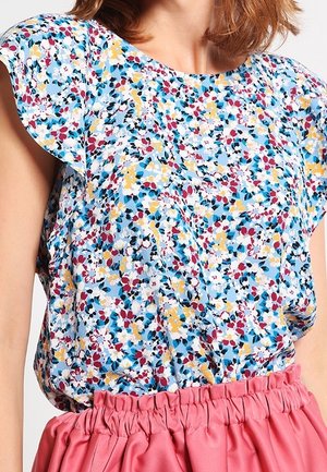 Kobieta w krótkim rękawie bluzki w kwiaty w niebieskie, żółte i czerwone wzory, w połączeniu z różową spódnicą z wysokim stanem.