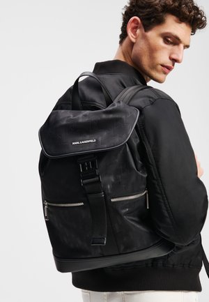 Black backpack featuring a textured design, front zip pocket, adjustable straps, and a flap closure with logo detail. Smooth nylon material.