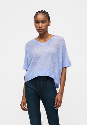 Moteris su pynėmis, apsirengusi šviesiai mėlynu laisvo mezgimo V formos iškirptės trumpomis rankovėmis megztiniu ir tamsiai mėlynomis džinsinėmis kelnėmis, ant balto fono.