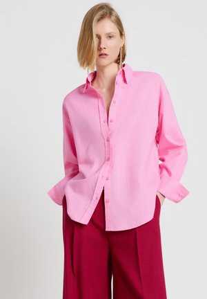 Frau mit kurzen blonden Haaren trägt ein lockeres rosa Hemd mit Knöpfen und hoch taillierte dunkelrote weite Hose, steht vor einem schlichten hellen Hintergrund.